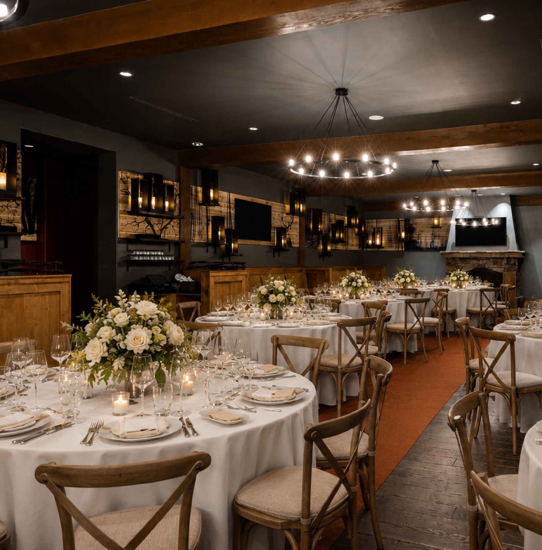 Elegant dining room with round tables set for an event, featuring white tablecloths, floral centerpieces, wooden chairs, wine glasses, and ambient lighting from chandeliers and wall sconces.
