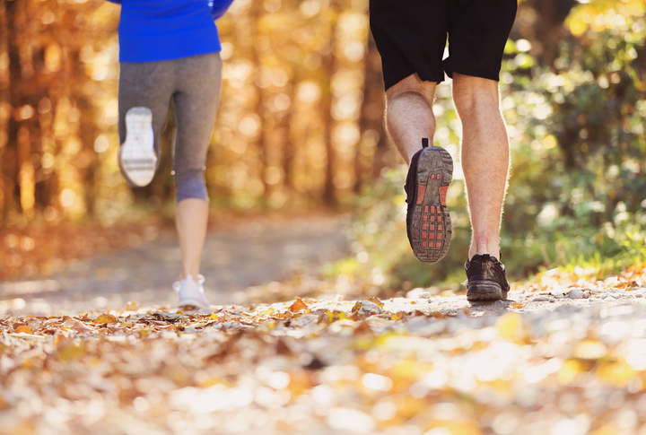 Two people jogging on a leaf-covered trail in a sunlit forest during autumn, with focus on their legs and running shoes.