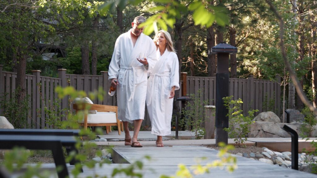 A man and woman in white robes walk together on a garden path, smiling and holding hands. Trees and greenery surround them, with a wooden fence and outdoor seating in the background.