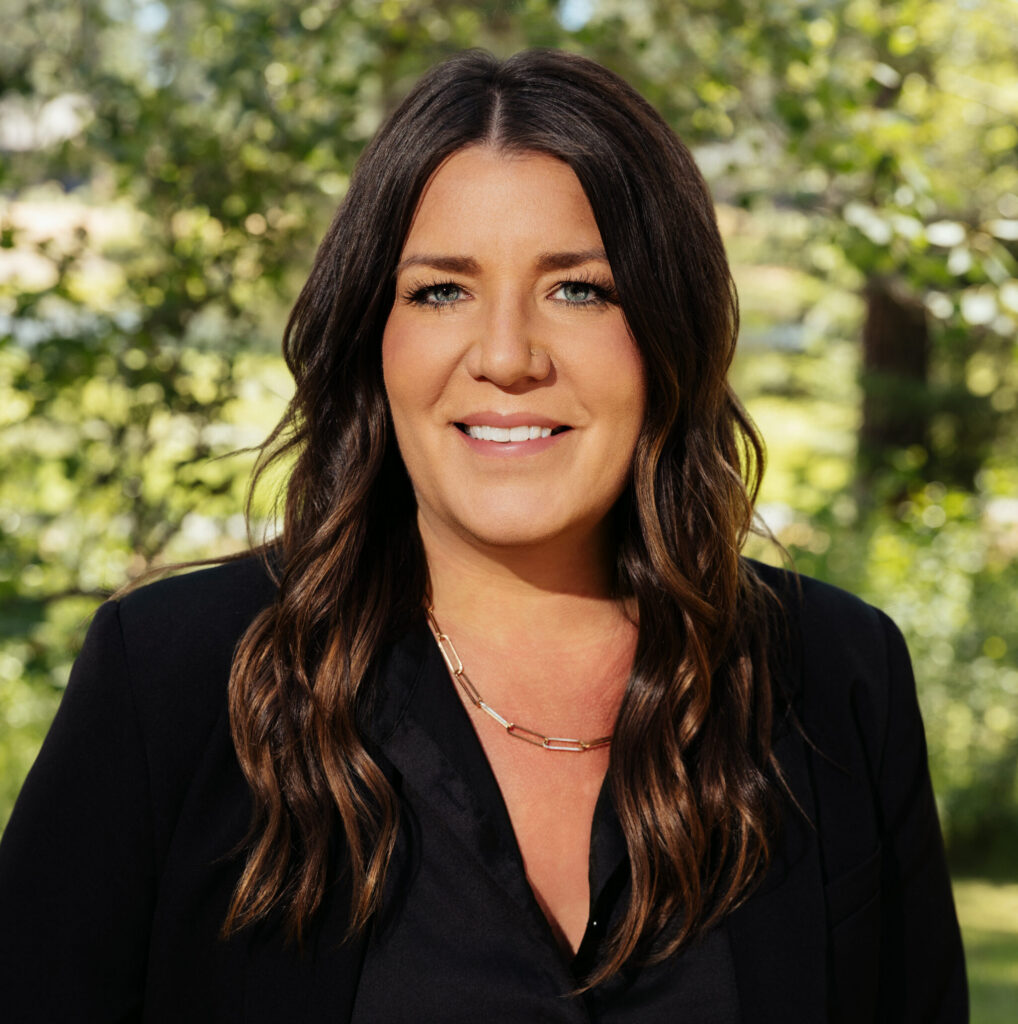 Woman with long brown hair wearing a black blazer and necklace, smiling outdoors with greenery and trees in the background.