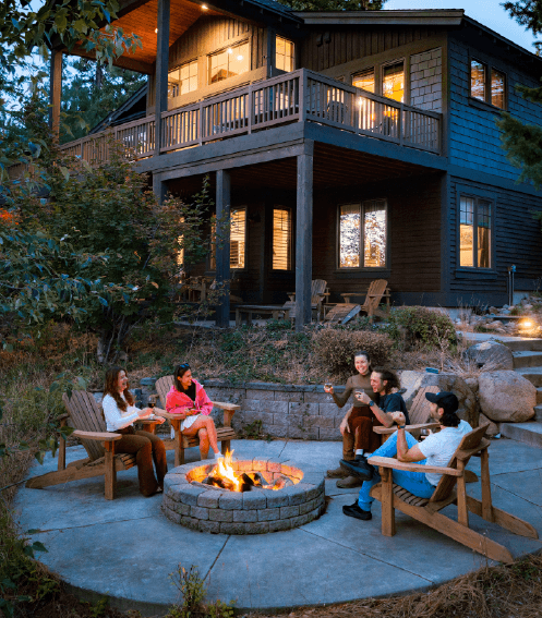 Four adults sit in wooden chairs around a stone fire pit on a patio in front of a two-story house at dusk.