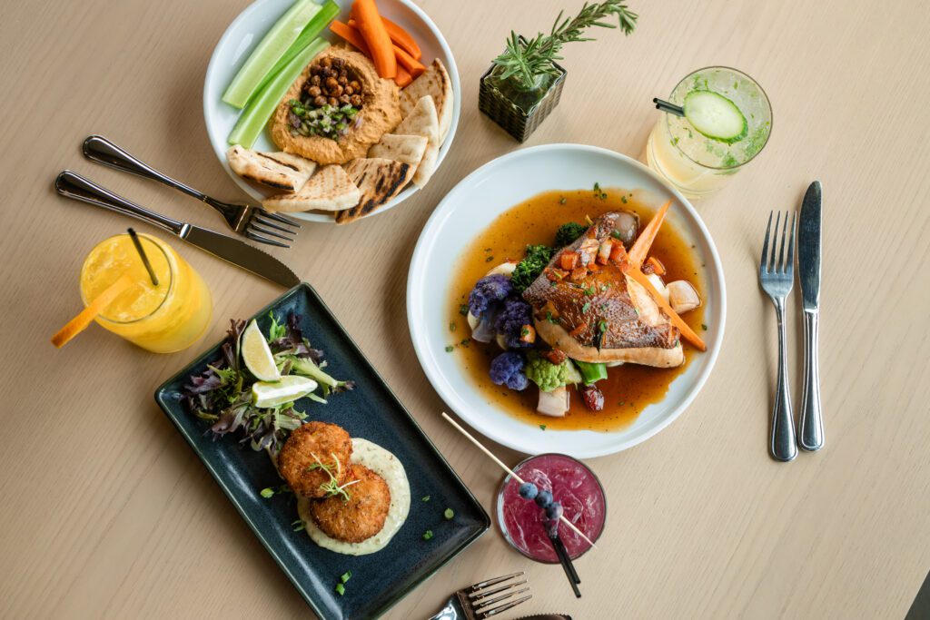 Three plates of food, including roasted chicken with vegetables, crab cakes with greens, and hummus with pita and vegetables, are arranged on a wooden table with three colorful drinks, utensils, and a small potted herb.