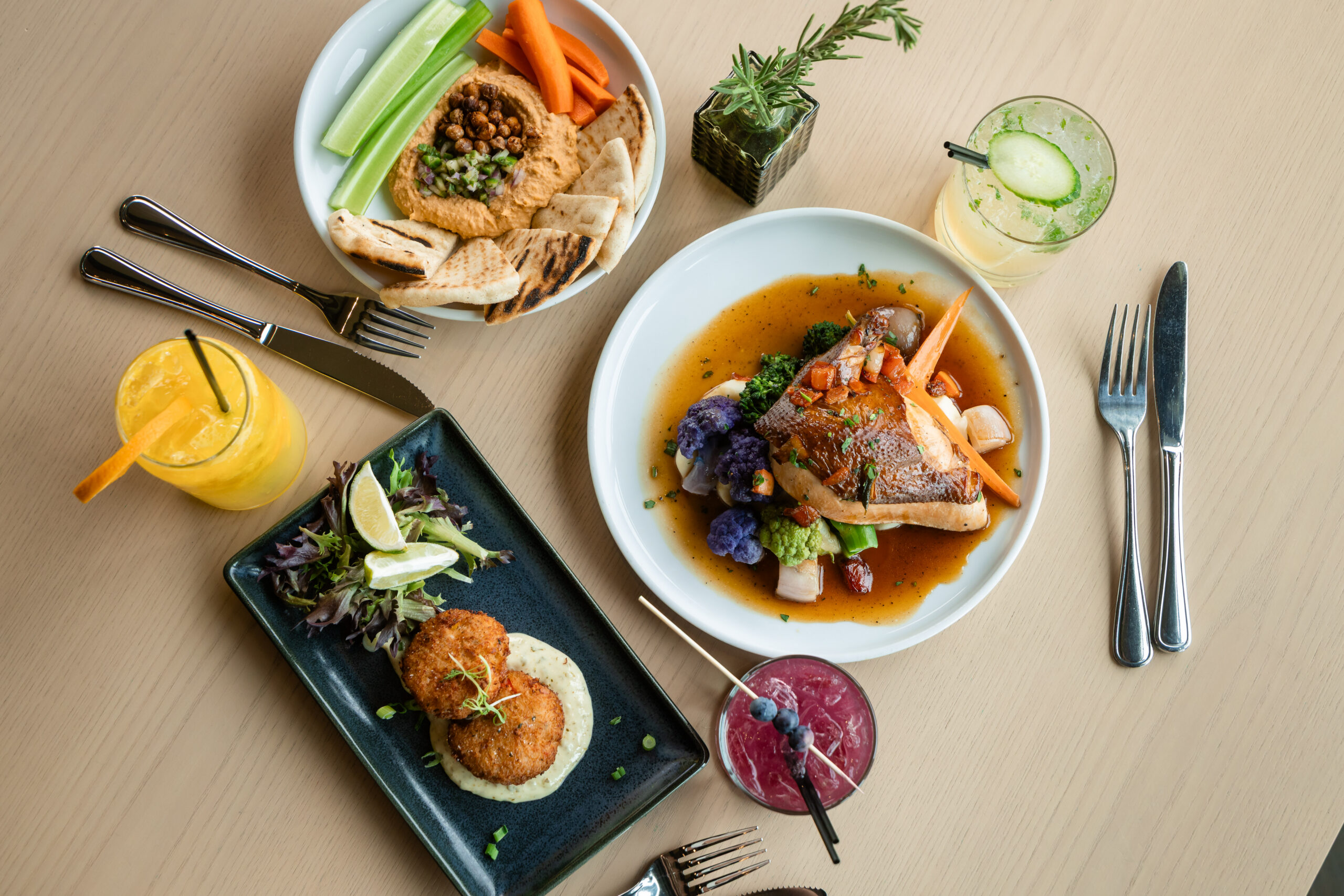 Three plates of food, including roasted chicken with vegetables, crab cakes with greens, and hummus with pita and vegetables, are arranged on a wooden table with three colorful drinks, utensils, and a small potted herb.