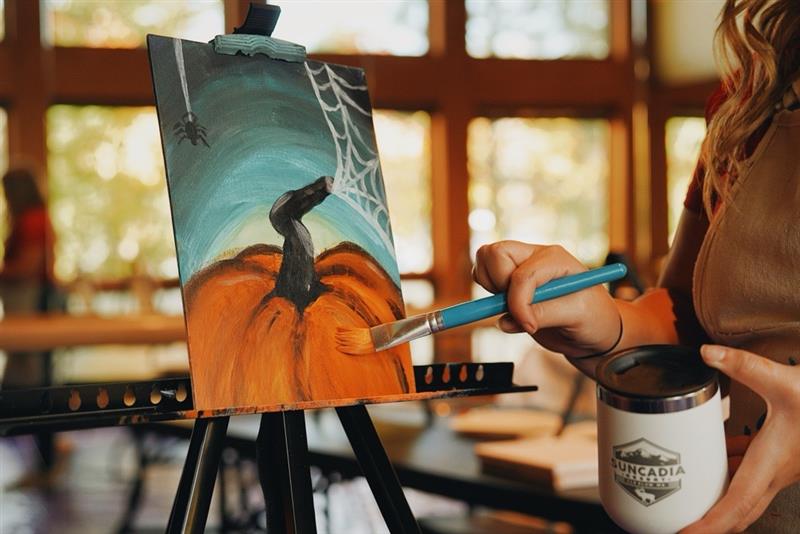 A person uses a paintbrush to add detail to a painting of a pumpkin on a canvas easel, while holding a Suncadia Resort tumbler.