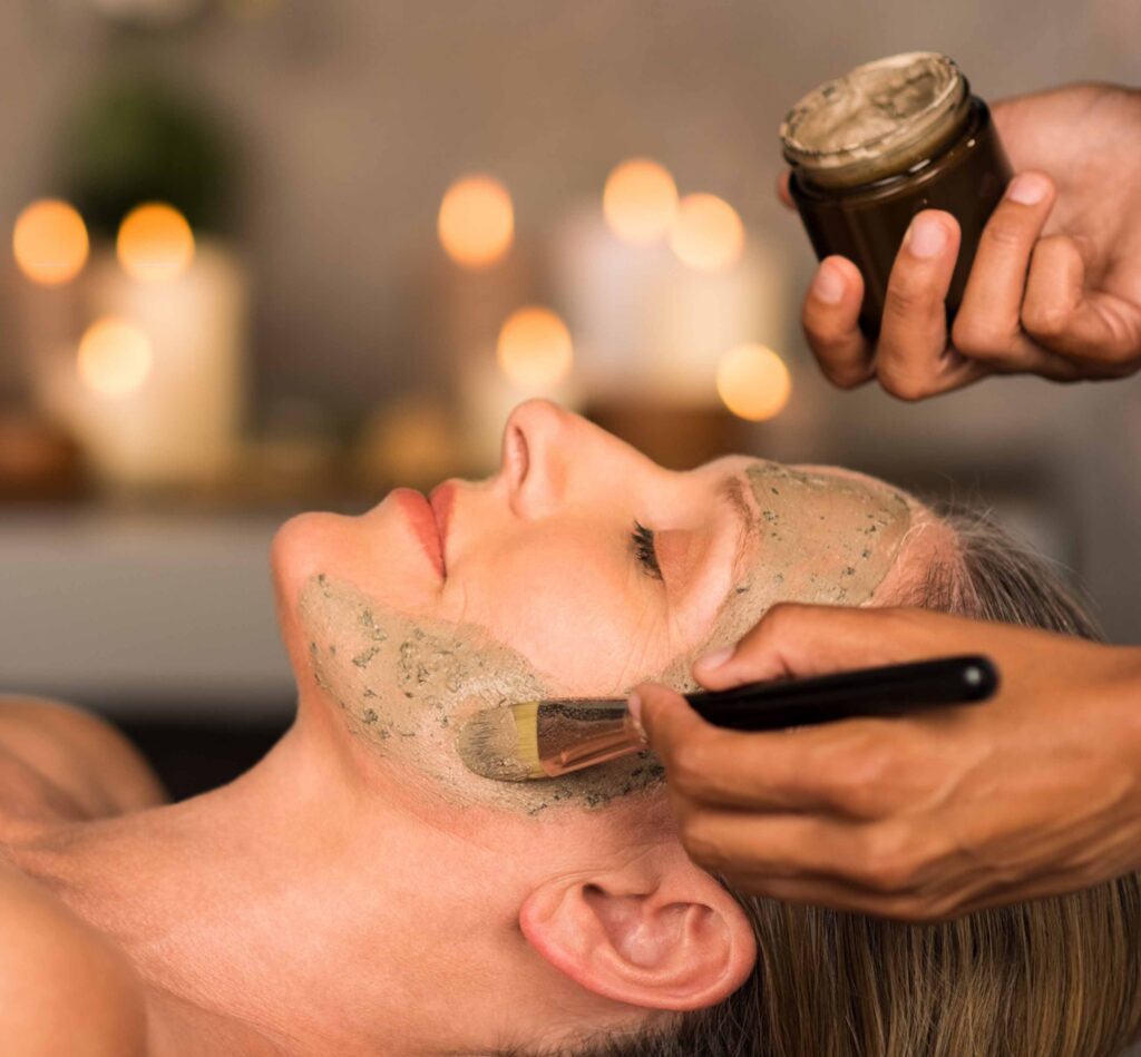 An aesthetician applies a mud mask to a womans face with a brush.